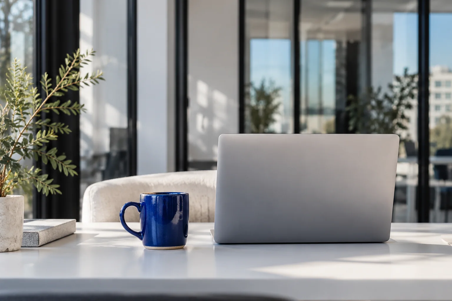 An inviting workspace with a laptop, cobalt blue cup, and sunlight through a glass partition