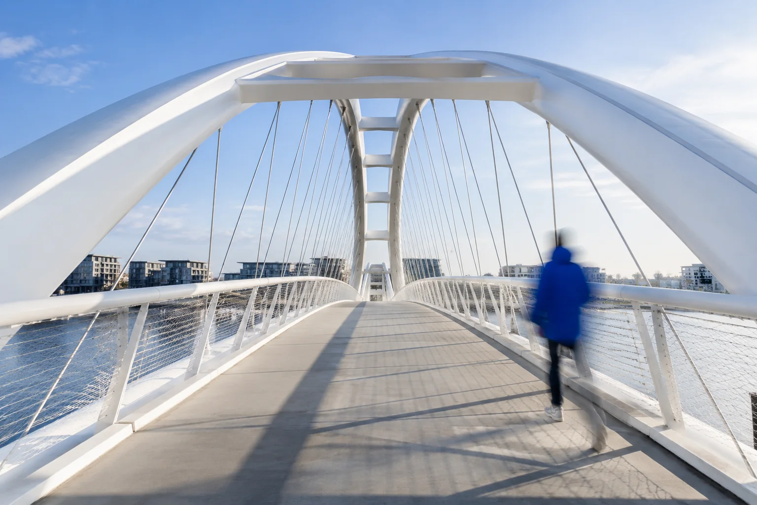 A modern white pedestrian bridge with one moving figure against a clear blue sky